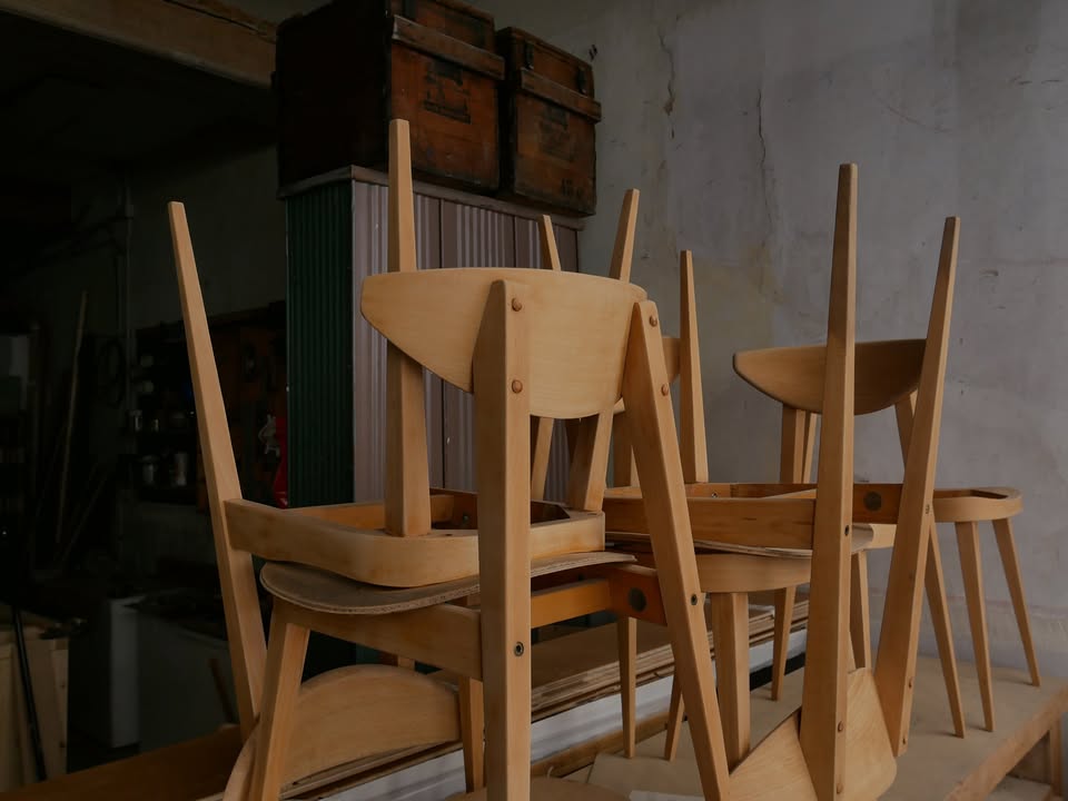 L'intérieur de l'atelier : des chaises en hêtre clair posées sur l'établi devant un mur de plâtre ancien, avec de vieilles caisses en bois et un meuble de rangement vert.